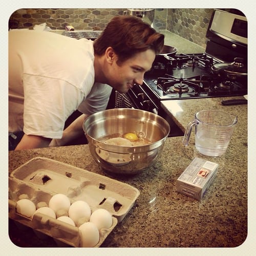 Cupcake ingredients and a mixing bowl with a man looking at a liquid measuring cup amount