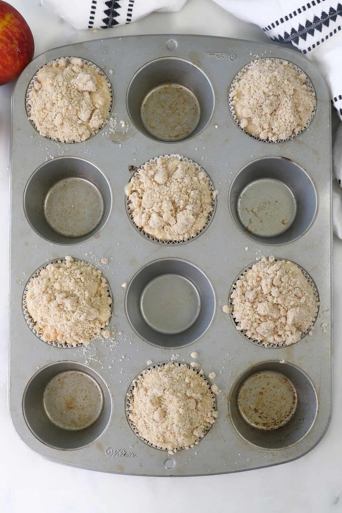 Overhead view of peach macadamia nut muffin batter in alternating wells of a muffin tin, topped with streusel.