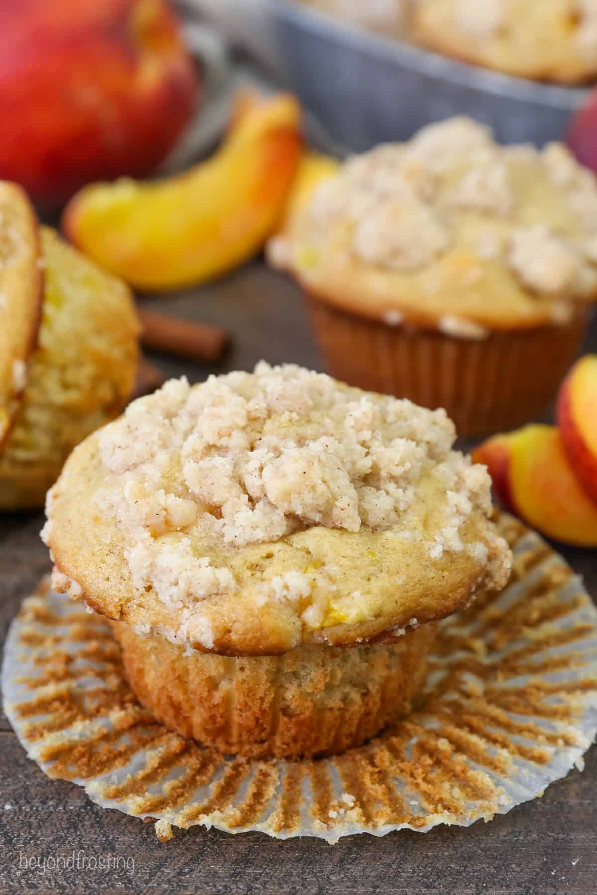 A peach macadamia nut muffin in an unwrapped muffin liner, with another muffin and peach slices in the background.