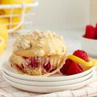 An almond raspberry muffin partially unwrapped from a muffin liner on a plate next to fresh rasbperries.