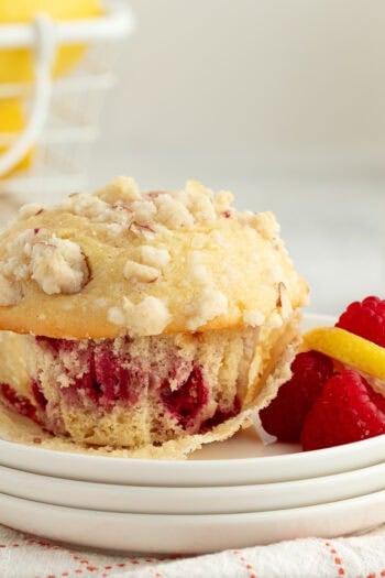 An almond raspberry muffin partially unwrapped from a muffin liner on a plate next to fresh rasbperries.