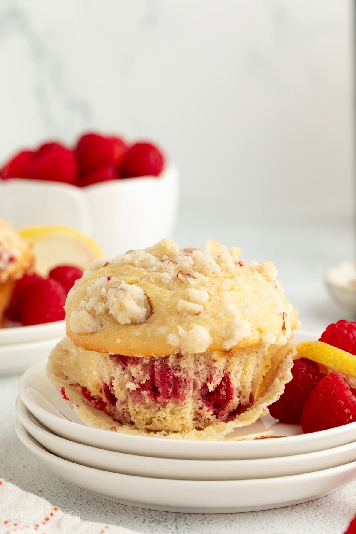 An almond raspberry muffin partially unwrapped from a muffin liner on a plate next to fresh rasbperries.