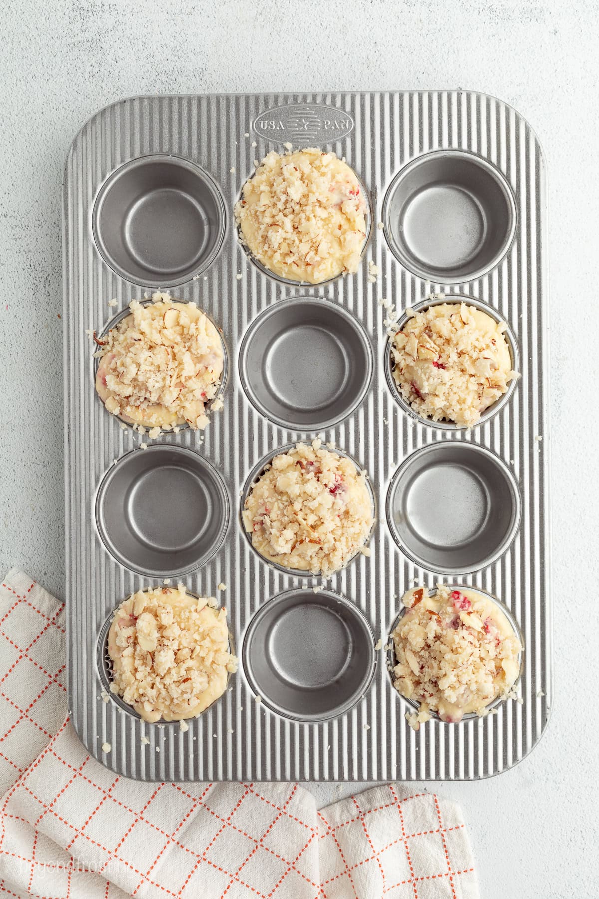 Overhead view of a muffin tin with every other well filled with raspberry muffin batter, topped with streusel.