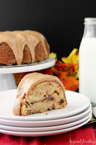 Slice of Apple Pie Coffee Cake with Maple Brown Butter Glaze on a plate in front of the whole cake on a cake stand