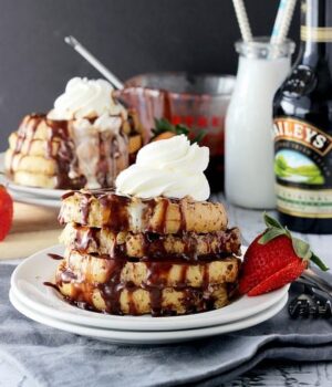 Bailey's Spiked French Toast stacked on a plate topped with chocolate drizzle and a dollop of whipped cream, with a strawberry on the side.
