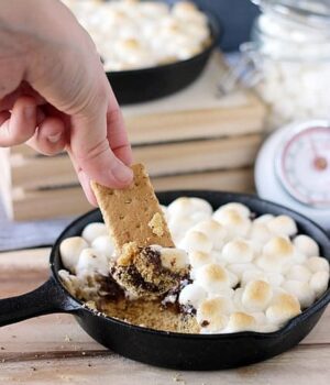 A skillet full of Cookie Dough S'mores Dip with toasted marshmallows, graham cracker crumbs, and melted chocolate being scooped up by a piece of a graham cracker.