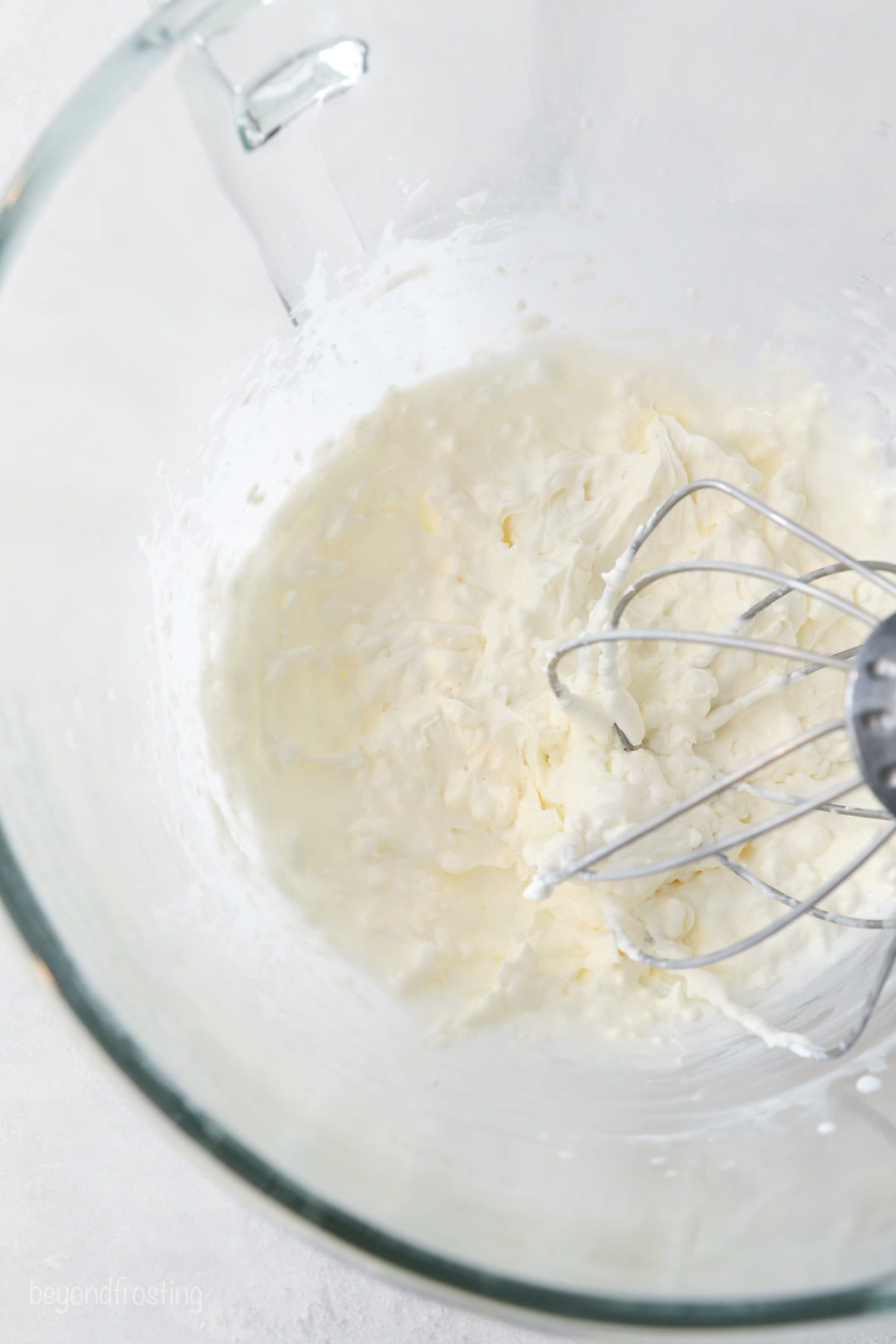 Beaten cream cheese in a glass bowl with a whisk attachment.