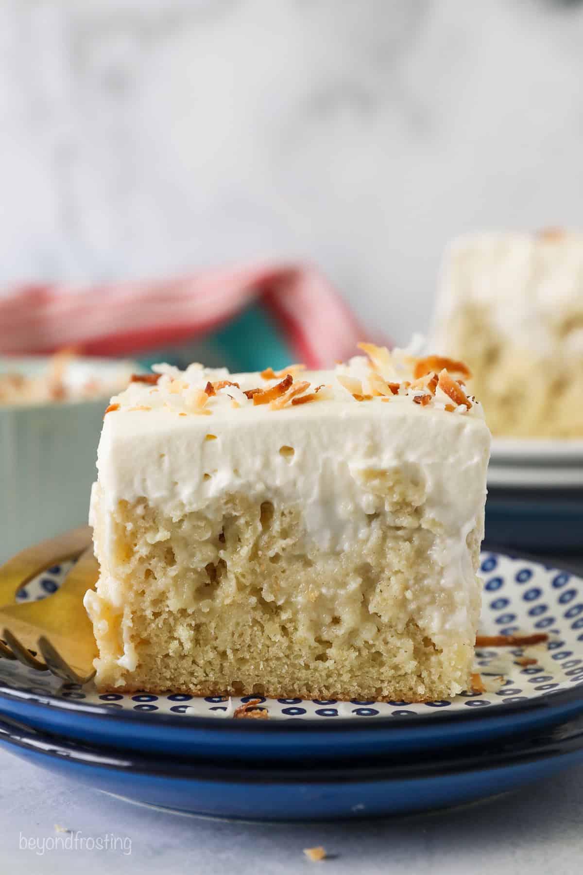 This coconut poke cake has soft, tender white cake drenched in creamy coconut filling, topped with cream cheese frosting and toasted coconut. Side view of a slice of frosted coconut poke cake topped with toasted coconut on a plate.