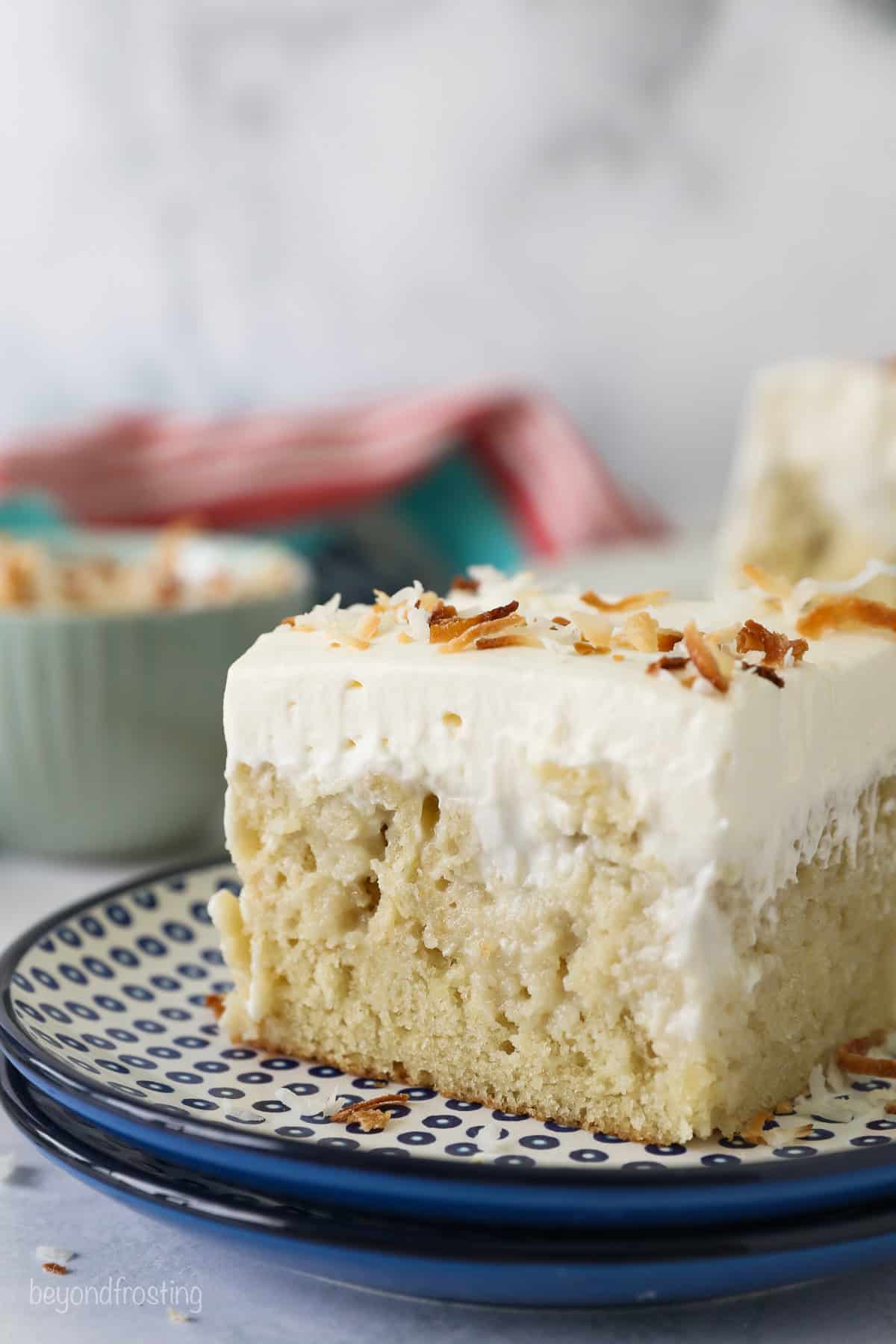 Side view of a slice of frosted coconut poke cake topped with toasted coconut on a plate.