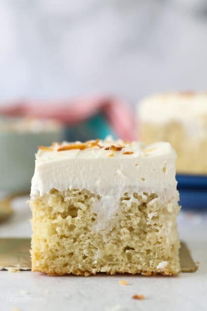 Side view of a slice of frosted coconut poke cake topped with toasted coconut.