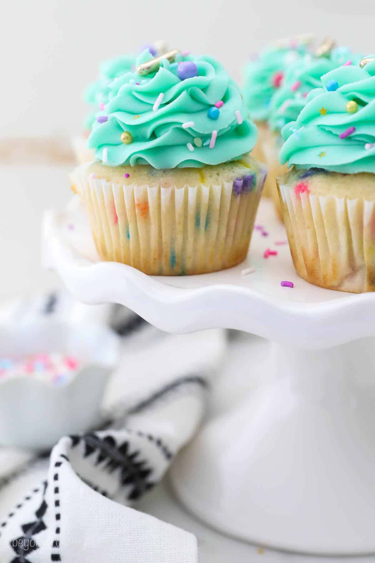 Funfetti cupcakes frosted with teal buttercream swirls and sprinkles on a white cake stand.