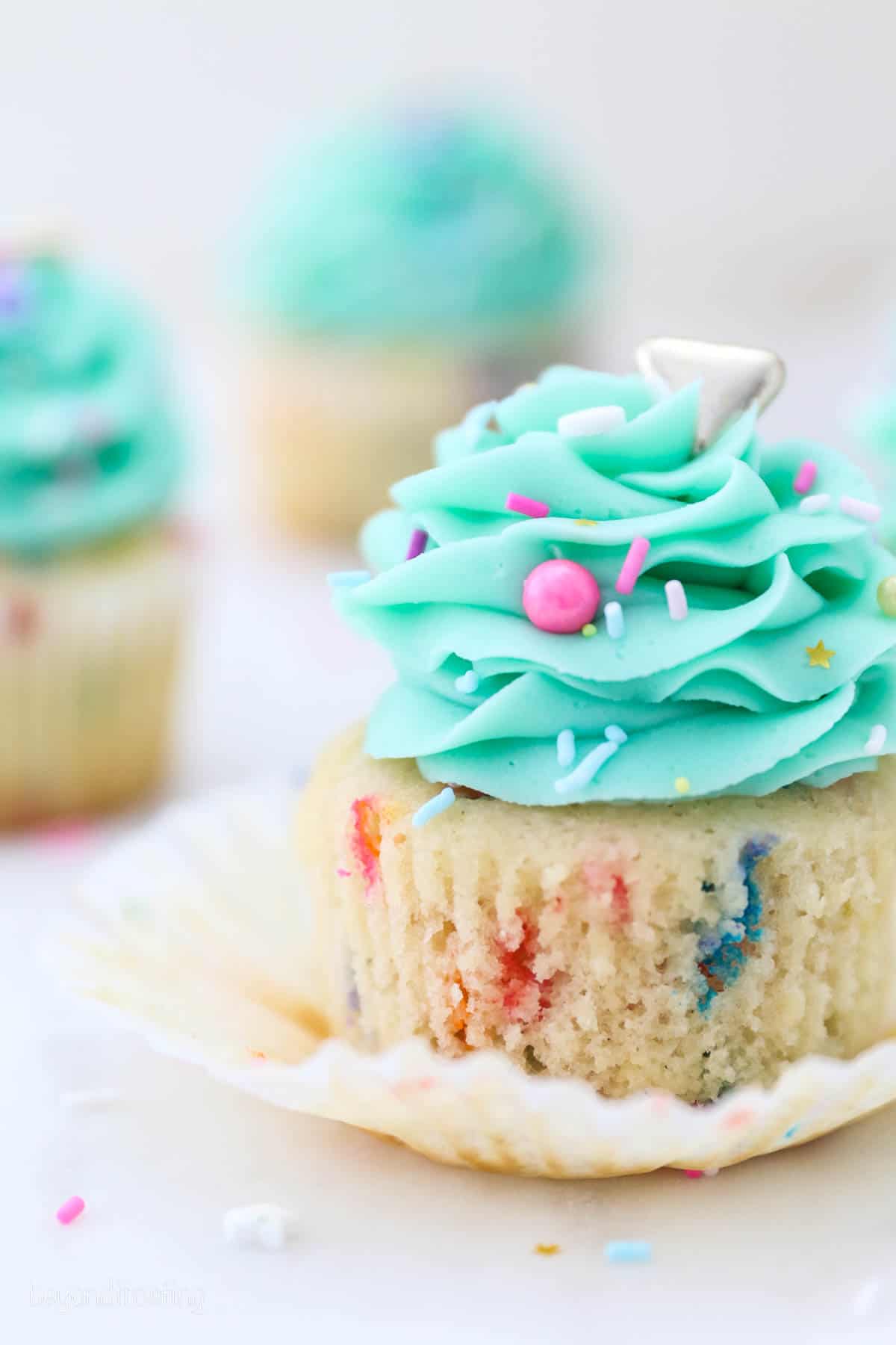 Close up of a partially unwrapped funfetti cupcake frosted with a teal buttercream swirl and sprinkles, with more cupcakes in the background.