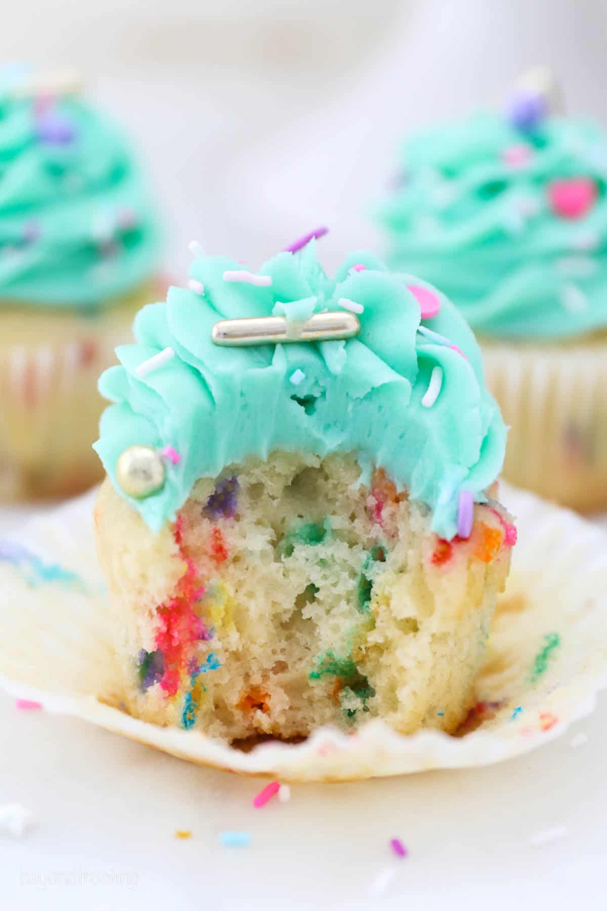 Close up of a partially unwrapped, frosted funfetti cupcake with a bite missing, with more cupcakes in the background.