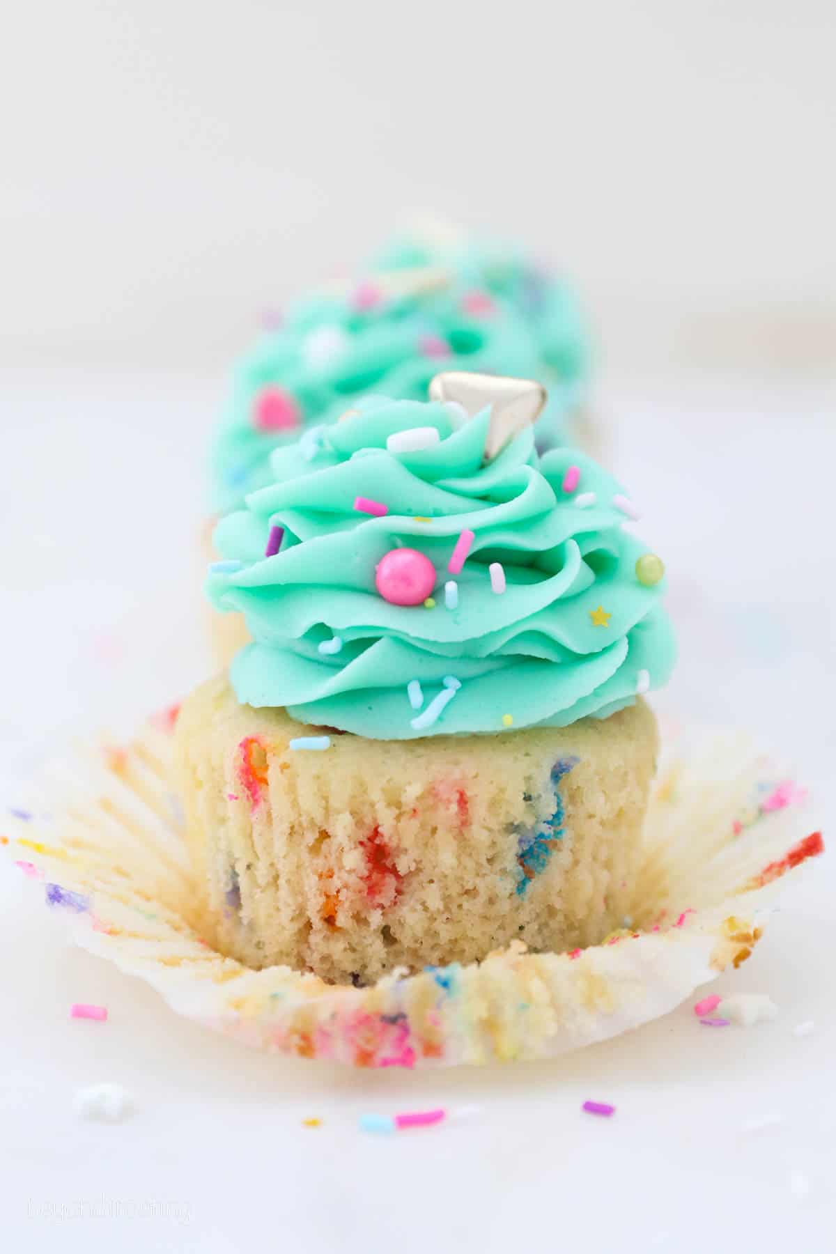 Close up of a partially unwrapped funfetti cupcake frosted with a teal buttercream swirl and sprinkles.