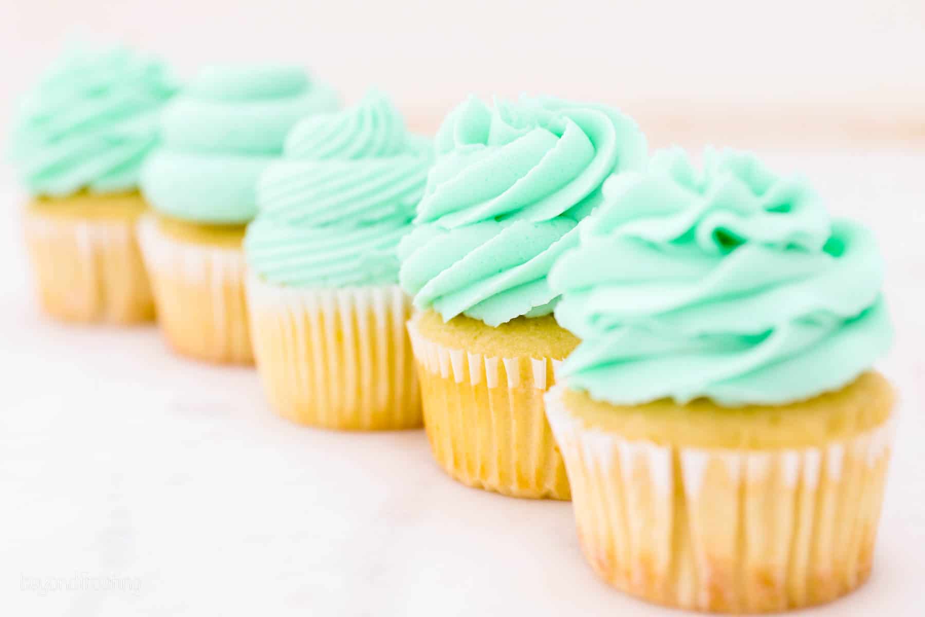 A row of vanilla cupcakes topped with swirls of teal frosting, piped with various piping tips.