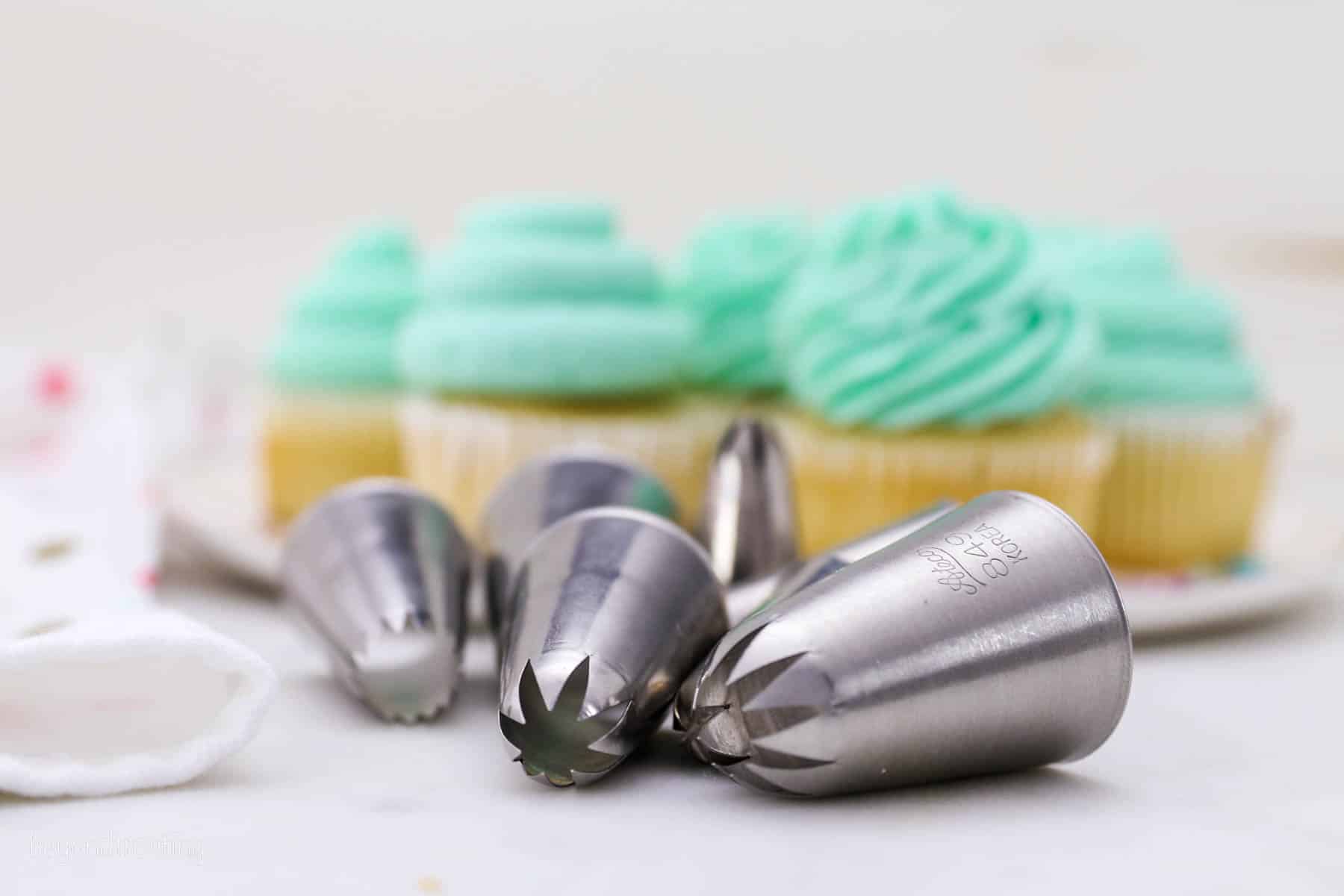 Assorted piping tips laying on a countertop in front of a tray of frosted vanilla cupcakes.