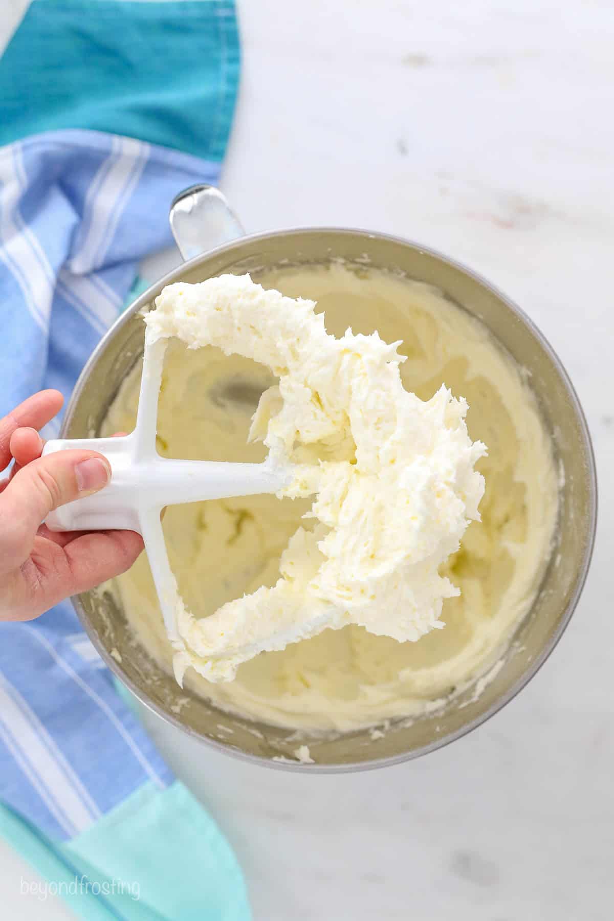 A hand holding a whisk attachment covered with vanilla frosting above a metal mixing bowl.