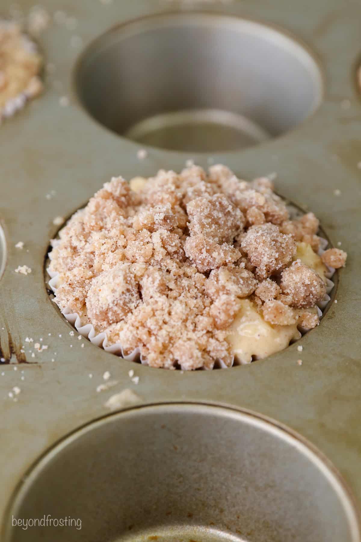 Close up of a well filled with apple muffin batter topped with streusel in a metal muffin tin.