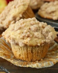 An apple muffin unwrapped from a paper liner on a countertop, with more muffins in a muffin tin in the background.