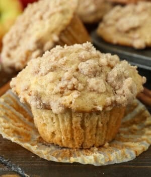 An apple muffin unwrapped from a paper liner on a countertop, with more muffins in a muffin tin in the background.