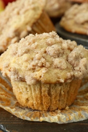 An apple muffin unwrapped from a paper liner on a countertop, with more muffins in a muffin tin in the background.