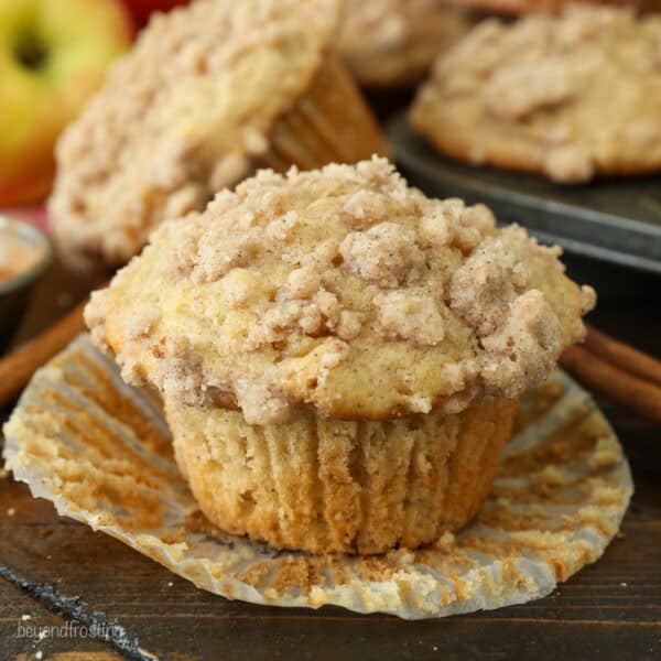 An apple muffin unwrapped from a paper liner on a countertop, with more muffins in a muffin tin in the background.