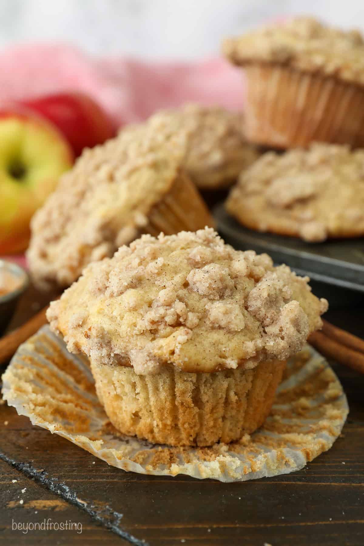 An apple muffin unwrapped from a paper liner on a countertop, with more muffins in a muffin tin in the background.