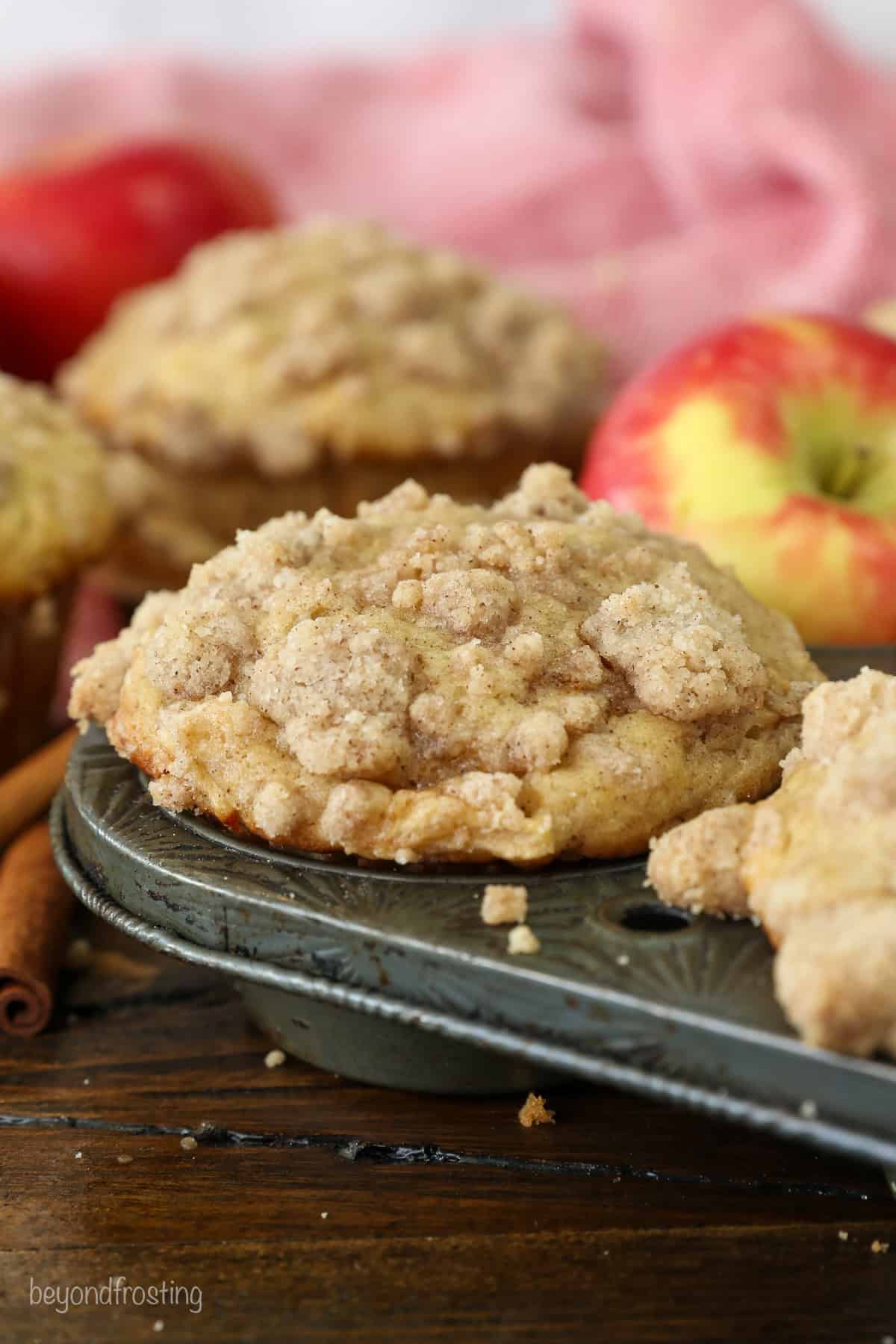 Close up of an apple muffin in the corner well of a muffin tin, with more muffins and whole red apples in the background.