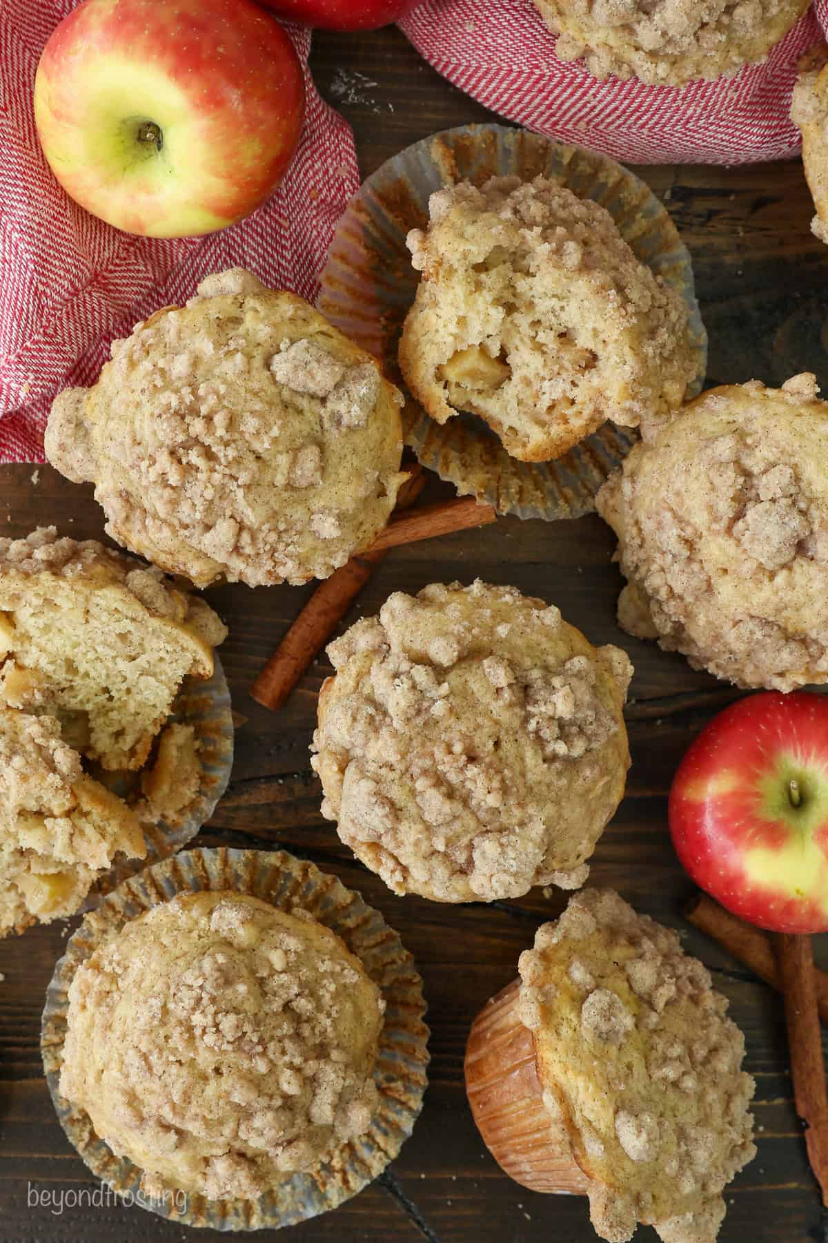 Overhead view of assorted apple muffins topped with cinnamon streusel, next to red apples on a countertop.