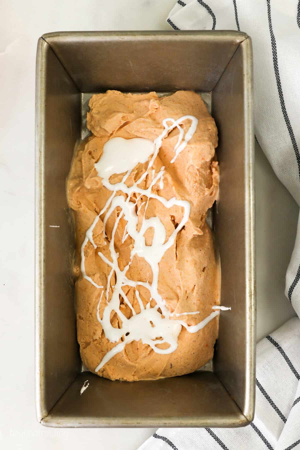 Melted marshmallow fluff swirled over sweet potato ice cream mixture inside a loaf pan.