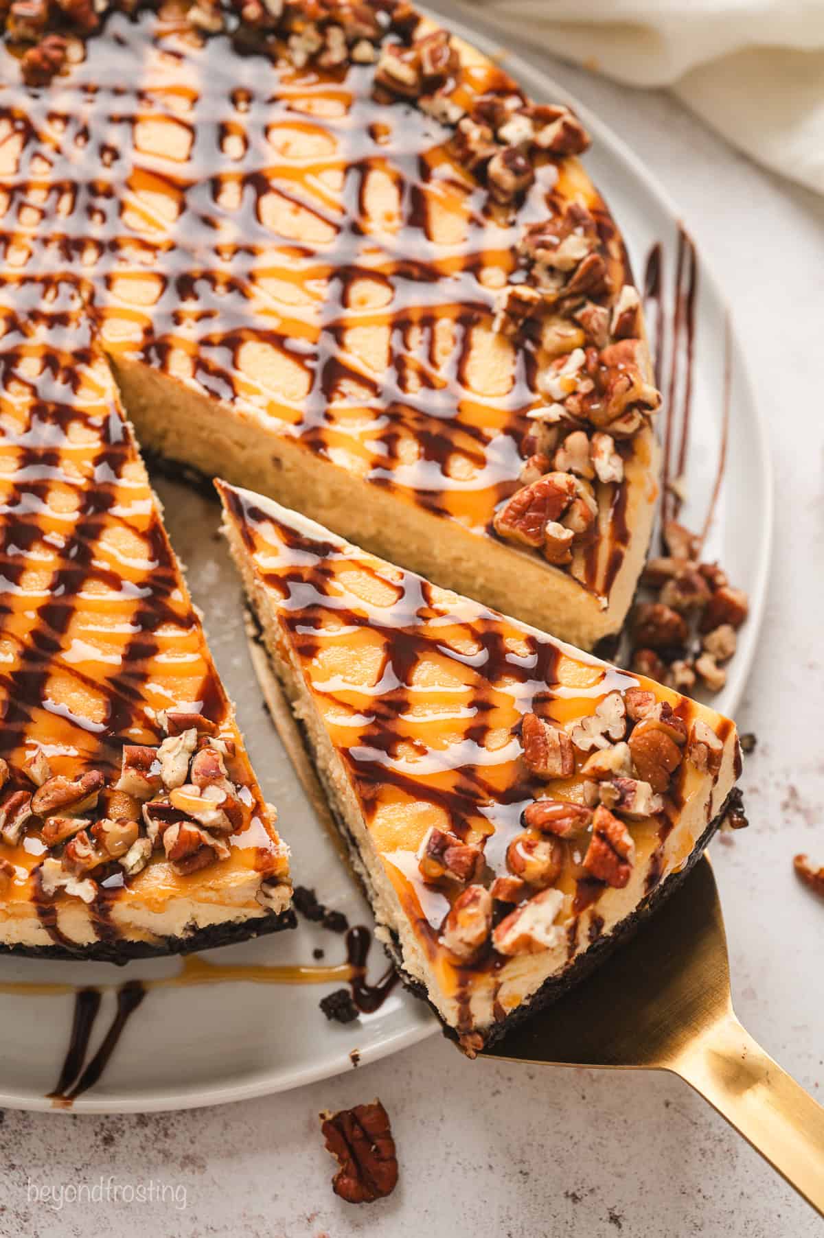 Overhead view of a slice of Turtle cheesecake being lifted from the rest of the cheesecake with a gold cake server.