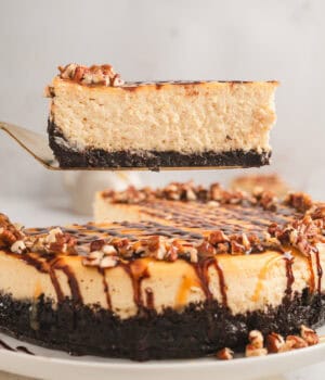 A slice of Turtle cheesecake being lifted from the rest of the cheesecake on a plate below.