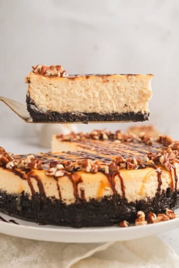 A slice of Turtle cheesecake being lifted from the rest of the cheesecake on a plate below.