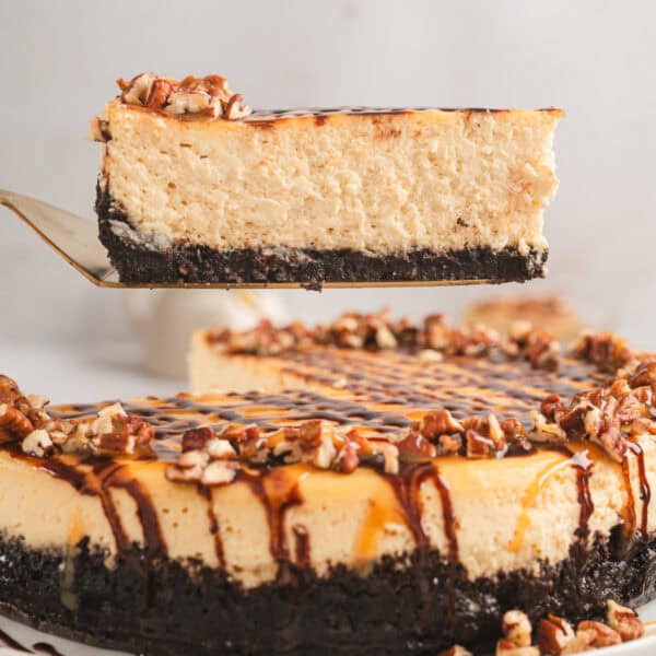 A slice of Turtle cheesecake being lifted from the rest of the cheesecake on a plate below.