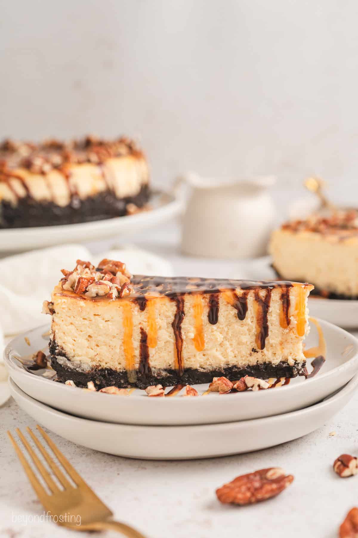 A slice of Turtle cheesecake on a plate with the rest of the cheesecake in the background.