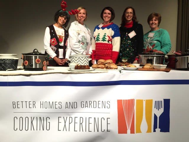 A group of food bloggers in holiday attire posing behind a demo table at BHG Cooking experience event