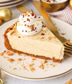 A slice of no-bake gingerbread cheesecake topped with whipped cream and Biscoff cookie crumbs on a white plate next to two forks.