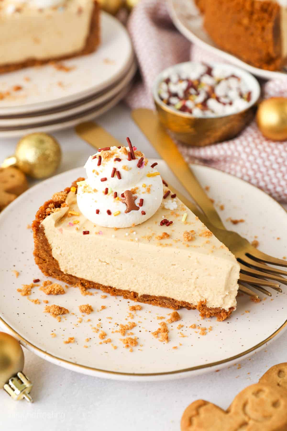 A slice of no-bake gingerbread cheesecake topped with whipped cream and Biscoff cookie crumbs on a white plate next to two forks.