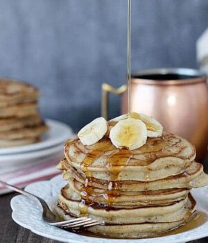 A stack of Peanut Butter Pancakes drizzled with maple syrup and topped with banana slices.
