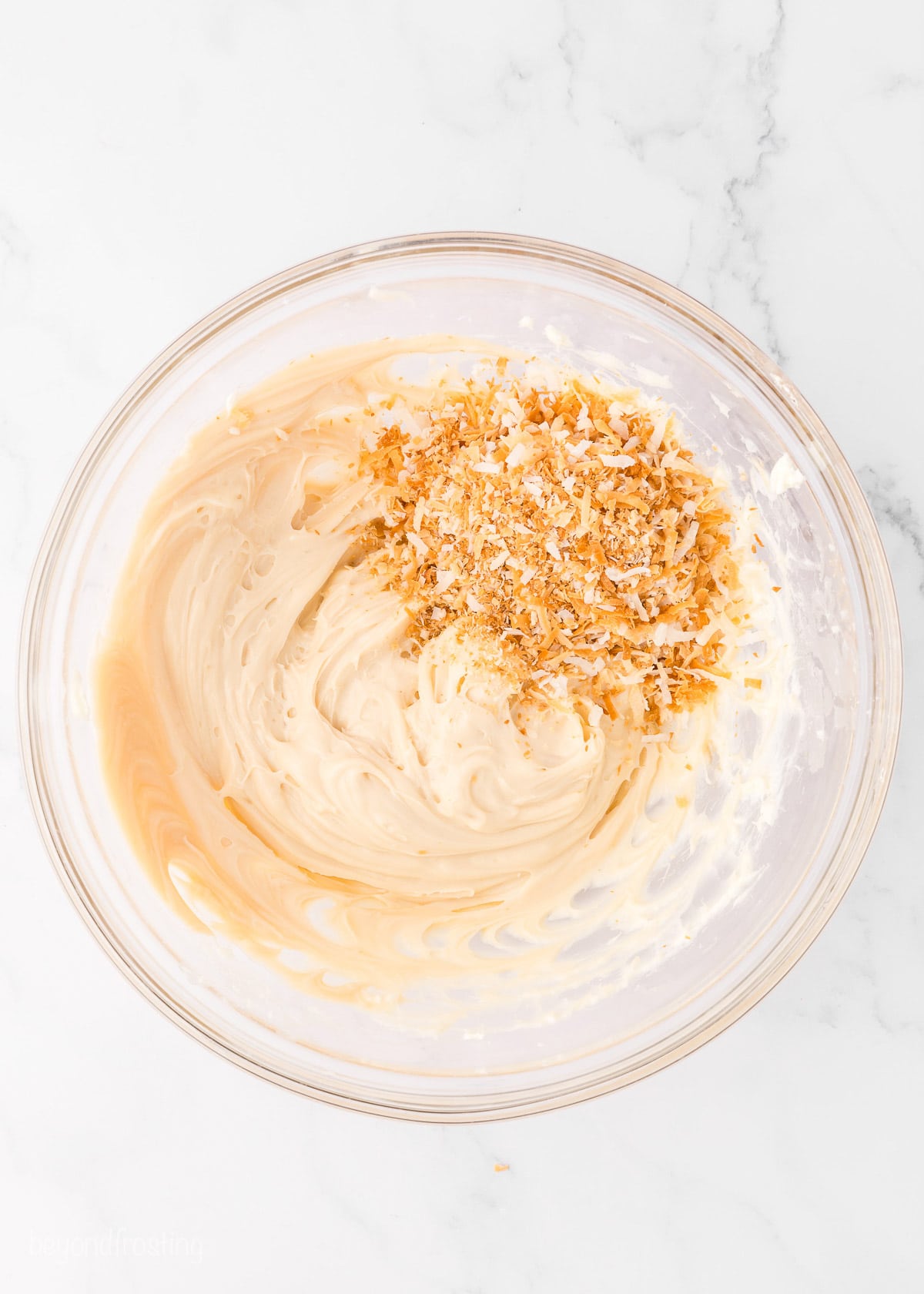 Toasted coconut added to a glass bowl with caramel cream cheese frosting.