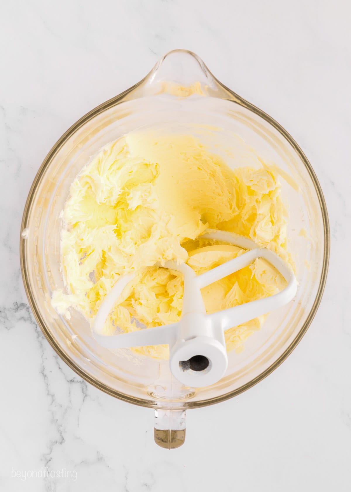 A stand mixer attachment resting in a bowl of whipped buttercream frosting ingredients.