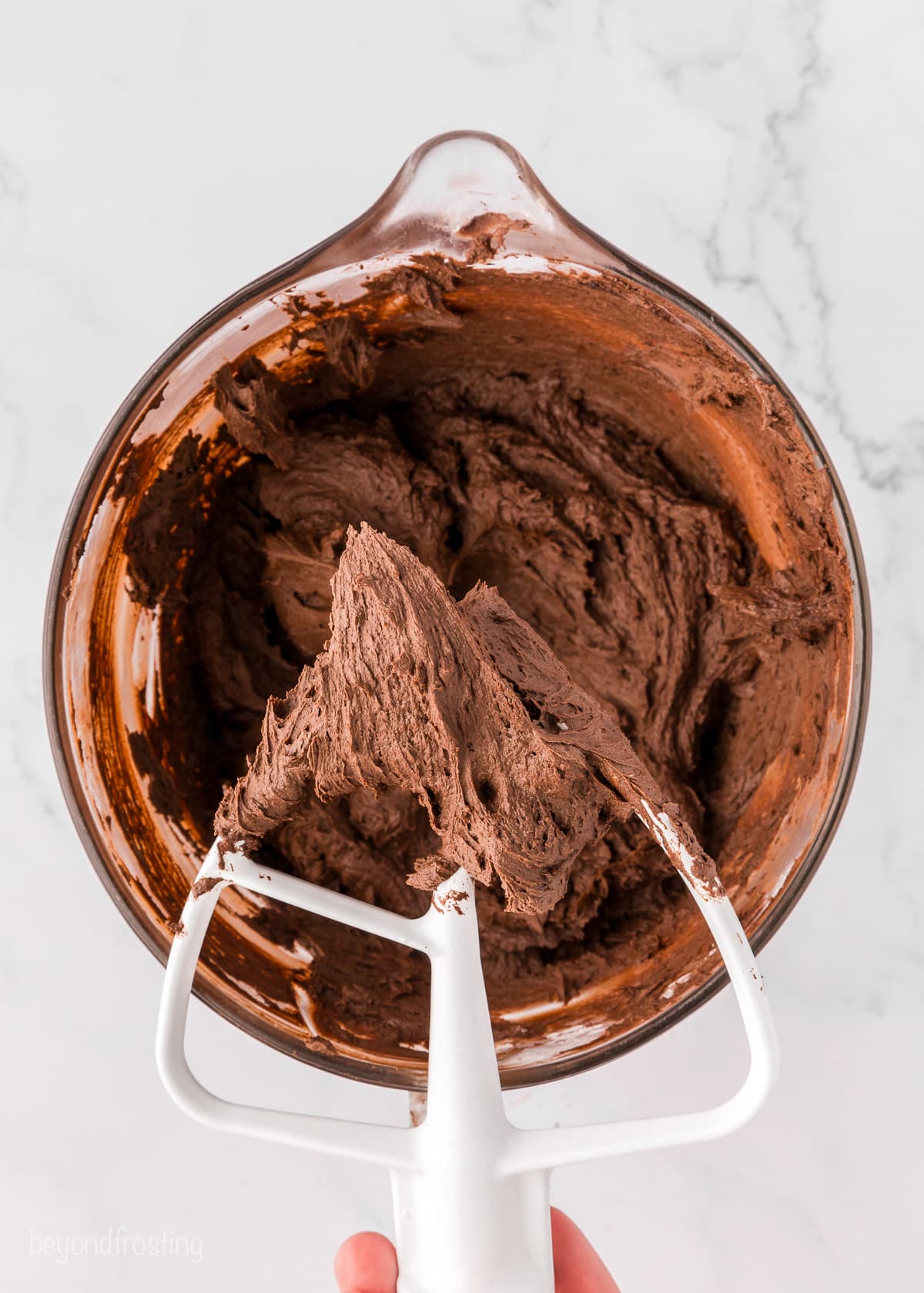 A hand holding a stand mixer attachment covered with chocolate buttercream over a mixing bowl filled with more frosting.