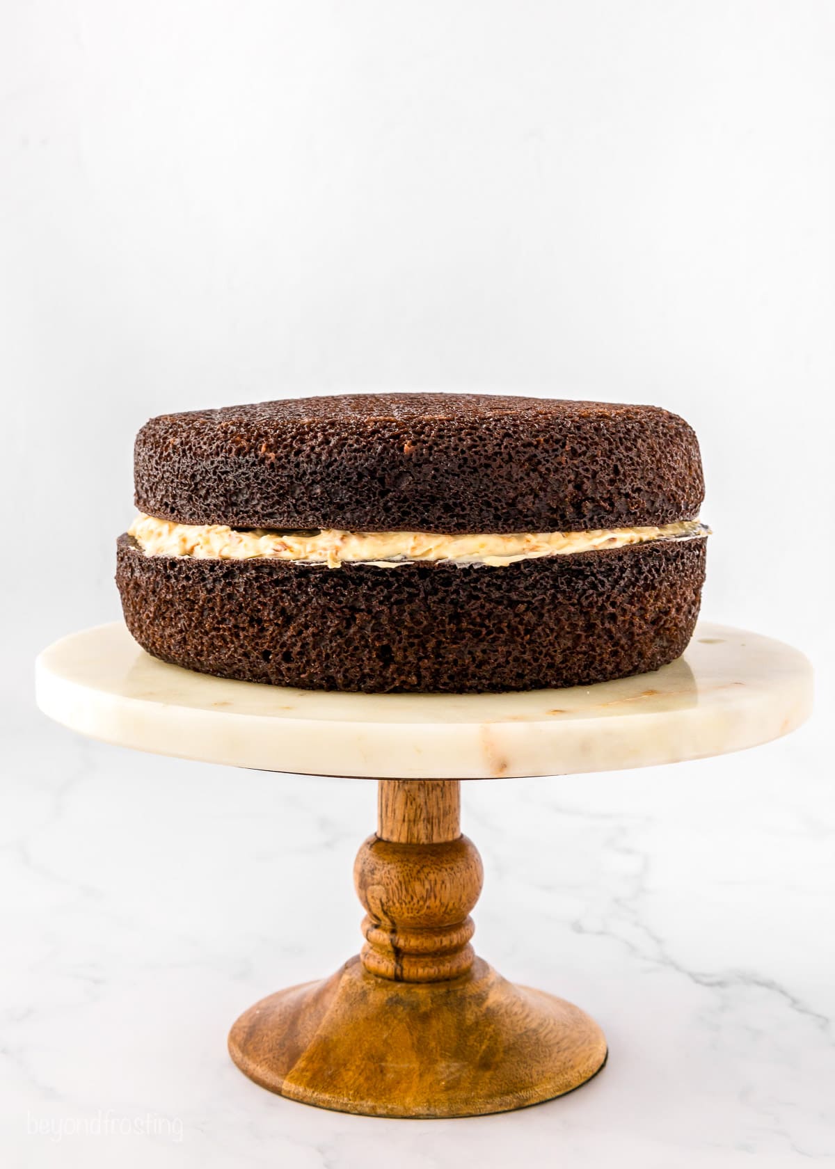 Two chocolate cake layers stacked and filled with caramel cream cheese frosting on a cake stand with a wooden base.