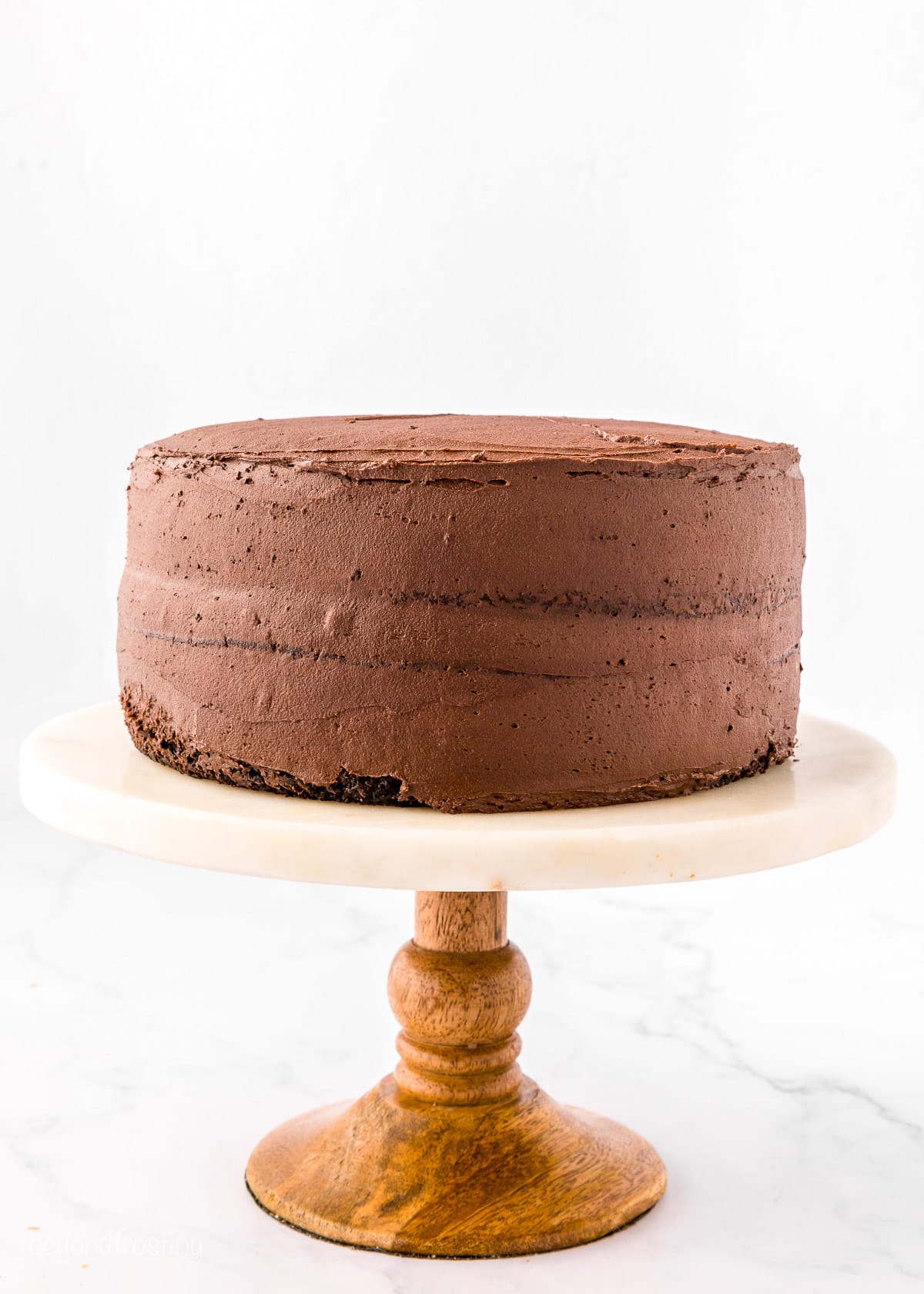 A two-layer chocolate cake frosted with chocolate frosting on a cake stand with a wooden base.