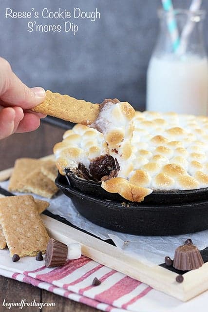 Ooey Gooey Reese's Cookie Dough S'mores Dip. The bottom layer is graham crackers, then it is topped with a peanut butter and chocolate cookie dough. This dessert dip is topped with toasted marshmallows. 