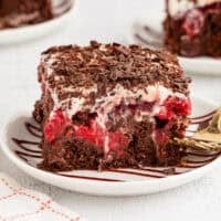 A slice of black forest poke cake next to a fork on a plate, with more cake slices on plates in the background.