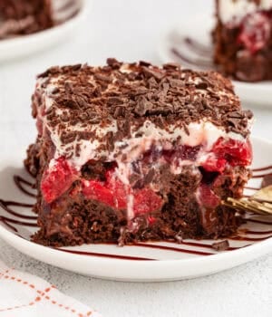 A slice of black forest poke cake next to a fork on a plate, with more cake slices on plates in the background.