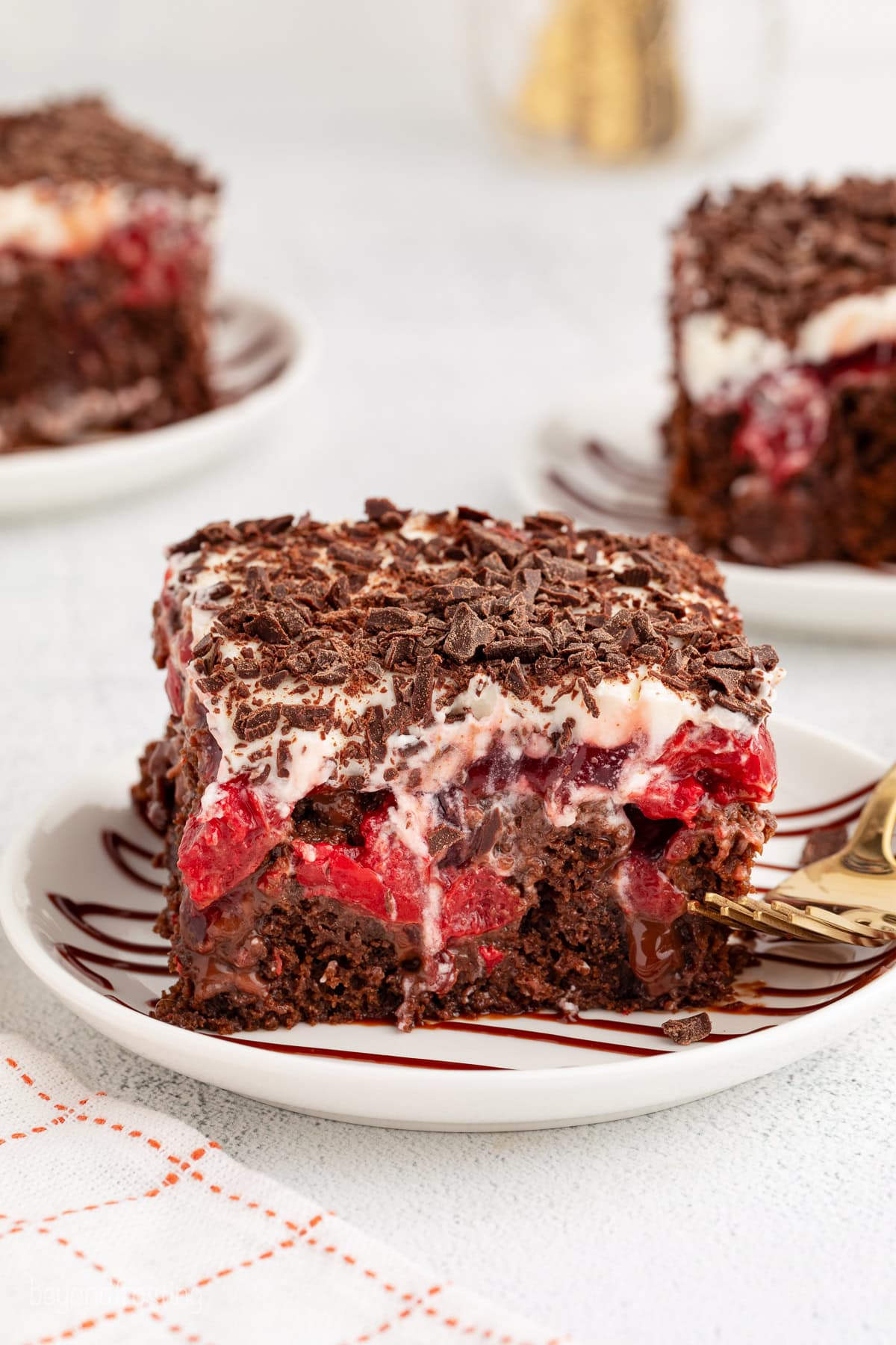 A slice of black forest poke cake next to a fork on a plate, with more cake slices on plates in the background.