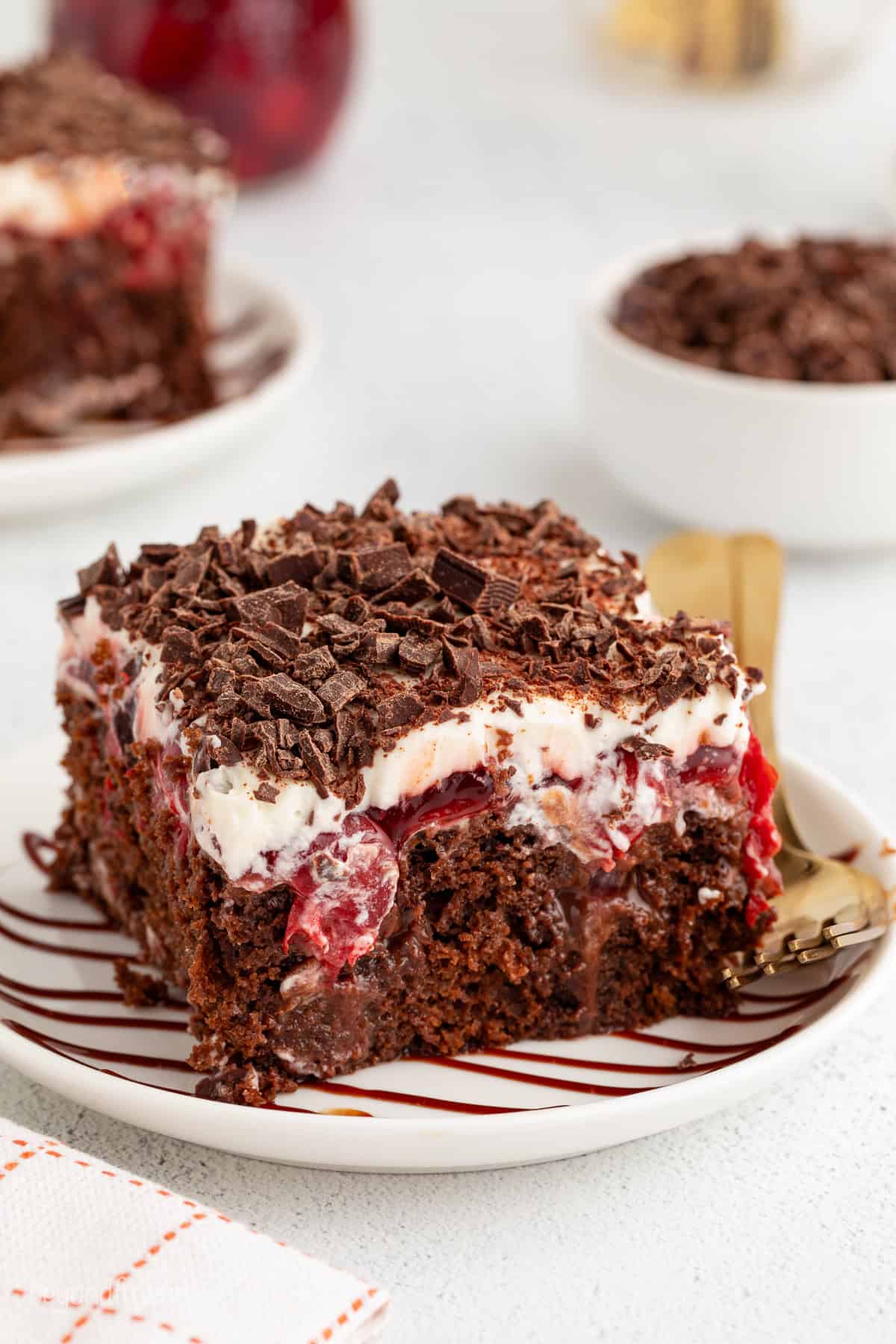 This easy black forest poke cake is a moist chocolate cake filled with hot fudge, topped with gooey cherry pie filling and whipped cream. A slice of black forest poke cake next to a fork on a plate.