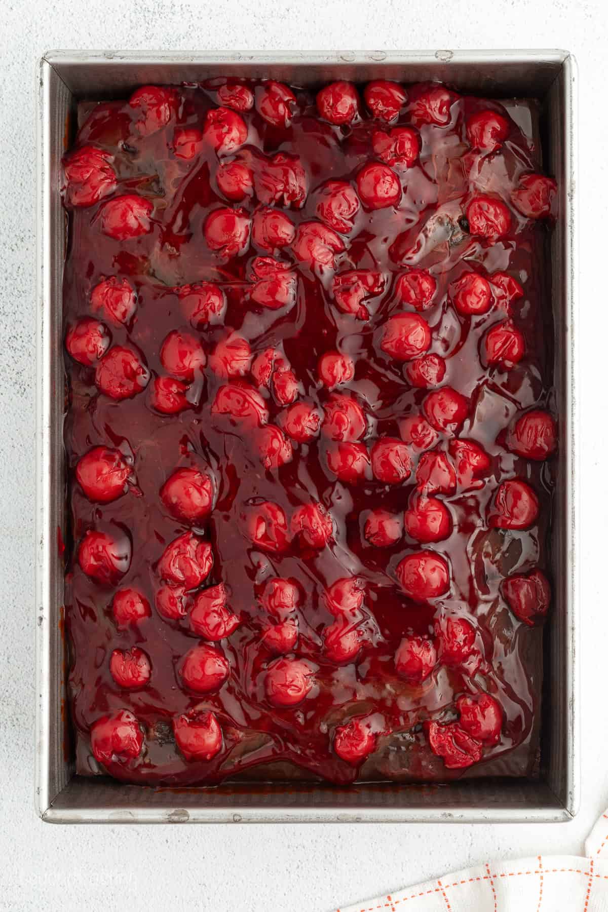 Overhead view of cherry pie filling spread overtop of a cooled chocolate cake in a 9x13-inch pan.